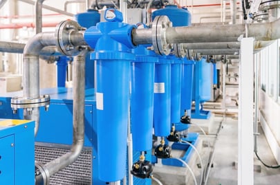 A close-up view of bright blue inline compressed air filters featuring a top-mounted pressure gauge and bottom drain valves. The filters are connected by stainless steel piping to a refrigerated air dryer unit in an industrial facility.
