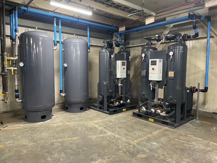 A modern industrial compressed air installation in a mechanical room, featuring two large grey receiver tanks on the left and two desiccant air dryers on the right, connected by blue aluminum piping
