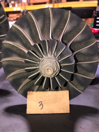 A dark, heavily worn and eroded centrifugal compressor impeller shown prior to restoration, sitting behind a small cardboard tag labeled '3'