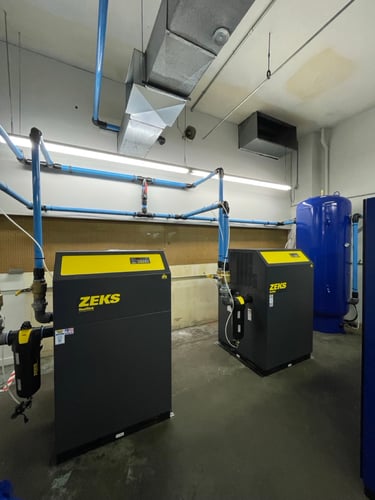 an industrial air compressor room with two dark grey and yellow ZEKS refrigerated air dryers and blue piping connected to an air storage tank