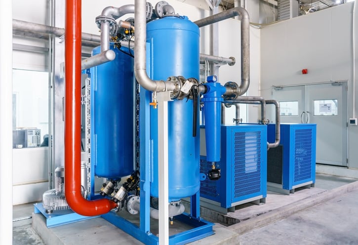 A comprehensive industrial compressed air treatment setup in a clean plant room. The system features large blue vertical tanks, interconnected stainless steel and red piping, an inline filter, and blue refrigerated air dryers in the background.