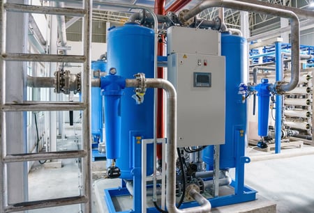 A large industrial twin-tower desiccant air dryer with two tall blue vertical tanks, complex metal piping, a central grey digital control panel, and smaller blue inline filters attached to the side intake and exhaust lines.