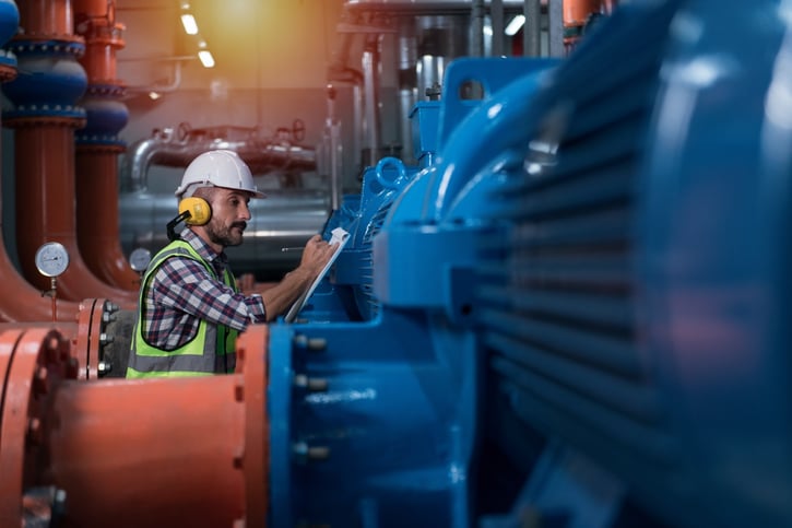 A professional maintenance technician wearing a hard hat, safety vest, and ear protection inspecting a large blue industrial air compressor unit and taking notes on a clipboard during an emergency service call.