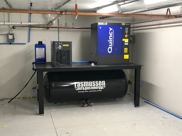 A compressed air system setup in a utility room, featuring a small black ZEKS refrigerated air dryer beside a blue Quincy QGS-15 air compressor, mounted on a metal table directly above a large, horizontal black Rasmussen Air & Gas Energy receiver tank.