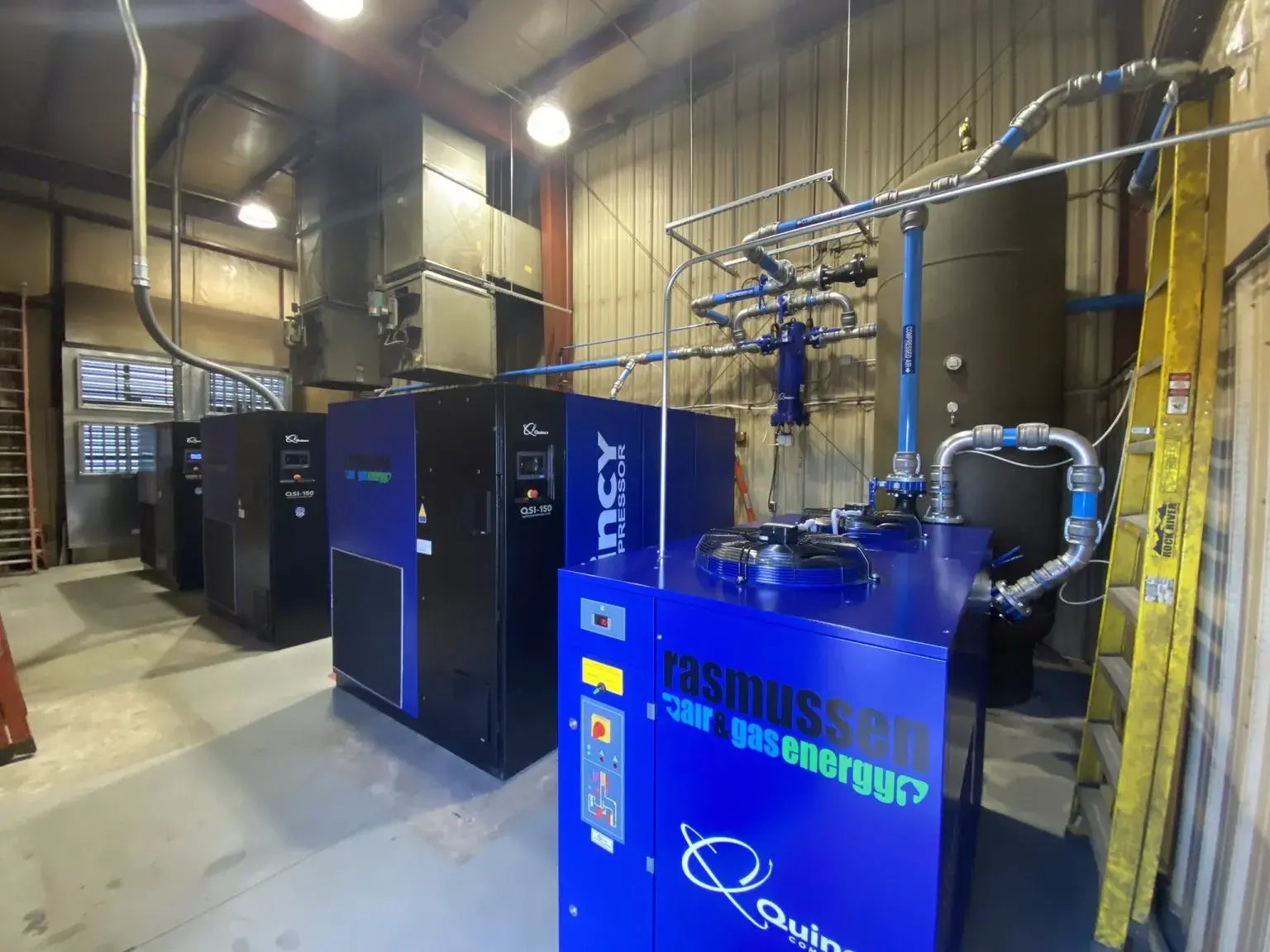 Industrial compressor room featuring Rasmussen-branded Quincy compressors and blue aluminum piping connected to air storage and filtration systems.