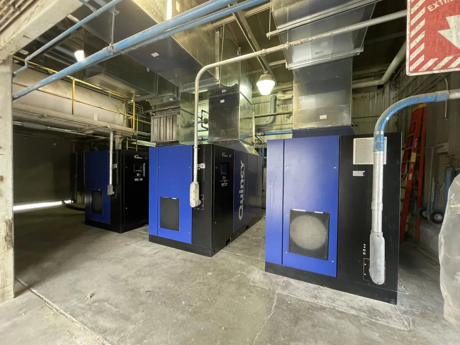 Three Quincy rotary screw air compressors installed in an industrial compressor room with connected ventilation and piping