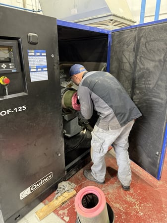 A Rasmussen Air and Gas Energy maintenance technician leaning into the open cabinet of a large black and blue Quincy industrial air compressor. He is holding a large, air filter with a bright pink end cap, while a matching filter sits on the red floor in the foreground.