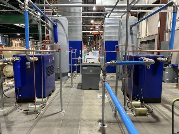 An integrated industrial compressed air system featuring blue air compressors, large vertical storage tanks, and overhead distribution piping in a manufacturing facility.