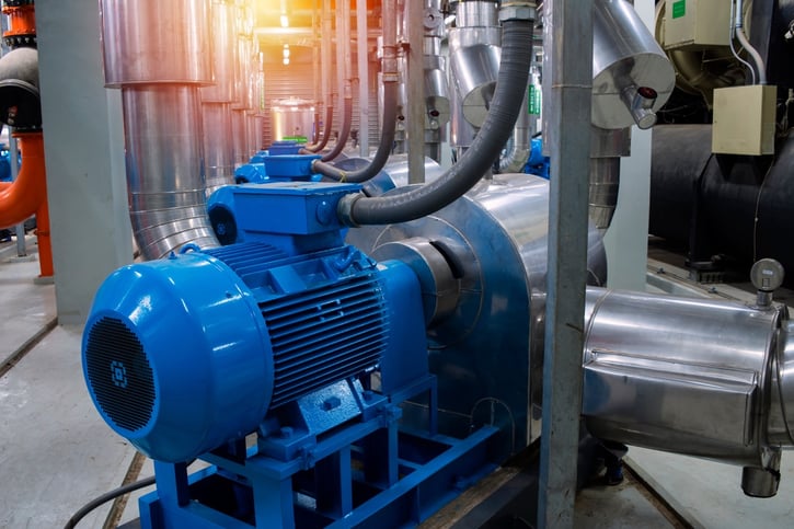 A large, bright blue industrial air compressor motor connected to heavy-duty insulated piping inside a factory, representing the type of commercial equipment that requires reliable cooling and maintenance during extreme summer heat.
